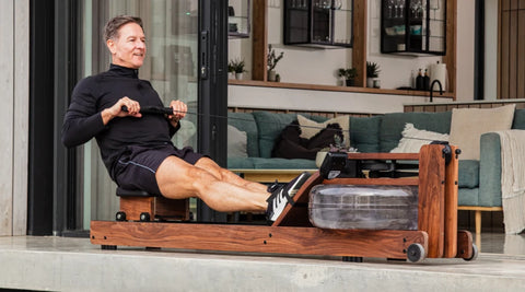 A man exercising on a WaterRower wooden rowing machine in a modern indoor living space with a green sofa and glass doors in the background.