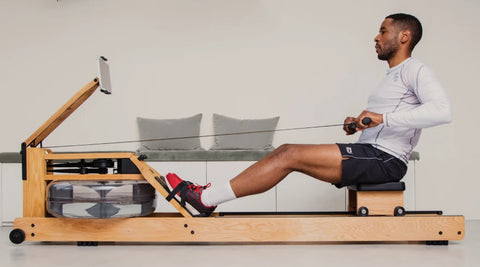 A man using a WaterRower Original wooden rowing machine in a minimalist indoor setting, wearing a white long-sleeve shirt, black shorts, and red sneakers.