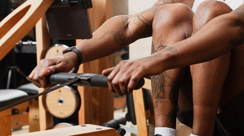 A close-up lifestyle shot of a person rowing on a WaterRower Original rowing machine, showing tattooed arms and legs, holding a black handlebar in a gym setting.