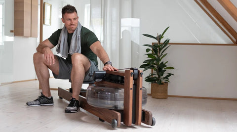 A man wearing a green t-shirt and grey shorts sits on a WaterRower Original wooden rowing machine in a bright, modern room with a potted plant in the background.