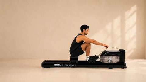 A man in a black athletic tank top and shorts is using a WaterRower Metallic exercise machine in a minimalist studio setting with warm beige walls and floor, with soft window light casting shadows.