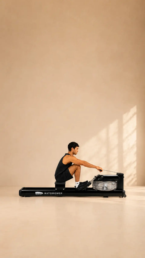 A man in a black athletic tank top and shorts is using a WaterRower Metallic exercise machine in a minimalist studio setting with warm beige walls and floor, with soft window light casting shadows.