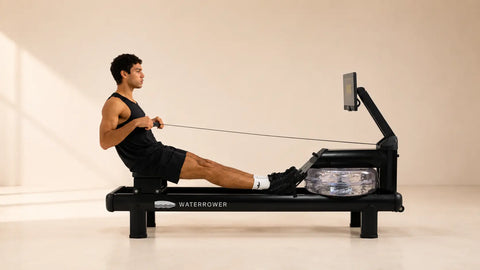 A man in a black athletic tank top and shorts is using a WaterRower rowing machine in a minimalist studio with warm beige walls and floor.