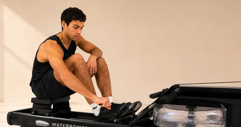A man in a black tank top and shorts sits on a WaterRower Metallic exercise machine, adjusting his shoe.