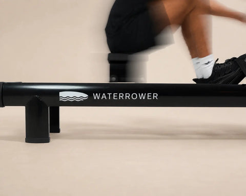 A close-up of a black metallic WaterRower with a person's legs and feet visible in motion on the upper deck against a beige studio background.