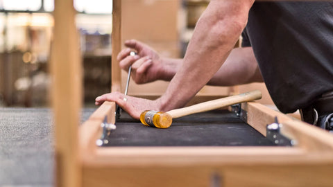 A close-up lifestyle shot of a craftsman's hands using a mallet and screwdriver on a wooden frame in a workshop setting.