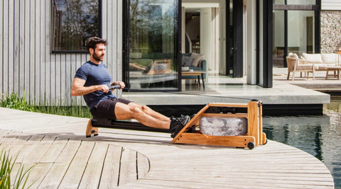 A man using a wooden WaterRower Hybrid rowing machine on an outdoor deck in front of a modern wooden house.