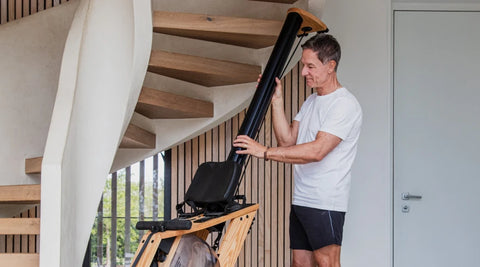 A man in a white t-shirt and black shorts standing a WaterRower Hybrid on it's end for storage inside a modern home with wooden stairs.