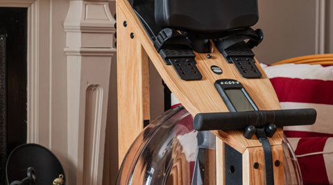 Close-up of a wooden WaterRower Hybrid Rowing Machine with black padded seat and handlebar, set in a home interior with a fireplace and striped cushion visible in the background.