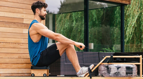 A man in a blue tank top and black shorts sits on a WaterRower Lite rowing machine outdoors next to a modern wooden building with large windows.