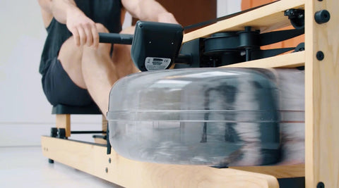 A person using a WaterRower Lite wooden water rowing machine, with the rowing handle, black motor unit, and large clear water tank in motion against a bright indoor background.