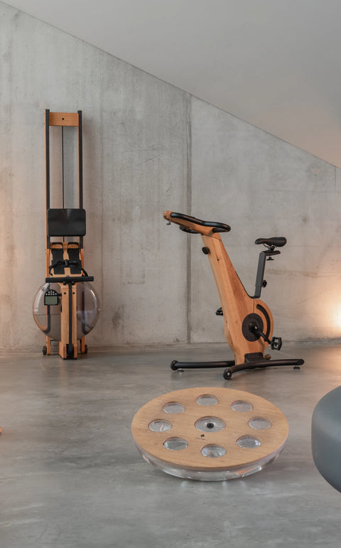 A warm-toned indoor gym scene with a wooden NOHRD Bike against a concrete wall, surrounded by NOHRD exercise equipment including SwingWeights, WaterRower Rowing Machine and an eau-me board. A large mid-century black fireplace occupies the right foreground.