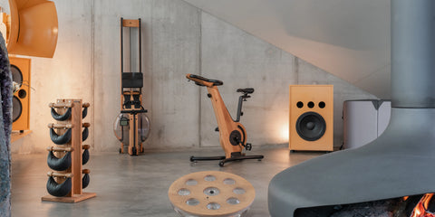 A warm-toned indoor gym scene with a wooden NOHRD Bike against a concrete wall, surrounded by NOHRD exercise equipment including SwingWeights, WaterRower Rowing Machine and an eau-me board. A large mid-century black fireplace occupies the right foreground.