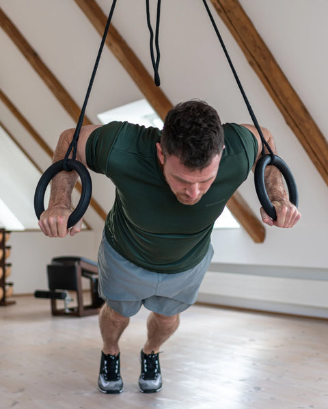 A man in a green t-shirt and grey shorts performing push-ups on a NOHRD Sling suspended from the ceiling of a loft-style room with wooden beams.