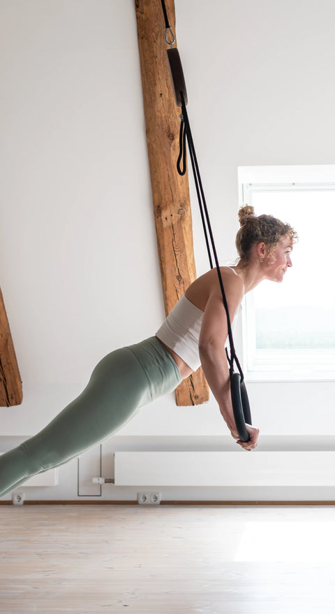 A woman in a home workout setting uses a NOHRD Sling to perform a push-up. She is wearing sage green leggings, a white tank top, and white trainers.