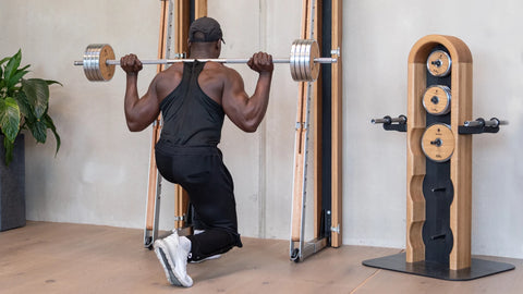 A man in a black sportswear performs a barbell knuckle exercise with a loaded barbell on his shoulders in a home gym setting, next to the wooden NOHRD Weight Plate Tower.