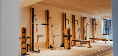 A fitness gym interior with multiple NOHRD SquatRacks against the wall with Weight Benches and Wood Dumbbells.