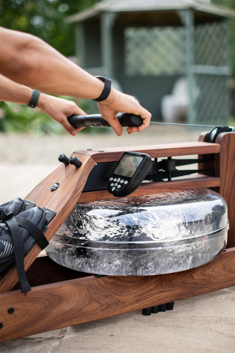 A close-up action shot of a person's arms and hands gripping the handle of the WaterRower Original. The rowing machine features a rich walnut wood frame, a large clear water-filled flywheel, and a black digital display screen.