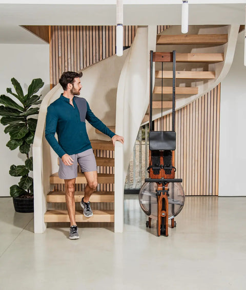 A man in athletic wear walks down a modern wooden staircase next to a WaterRower Original standing on it's end, set in a bright, contemporary home interior.
