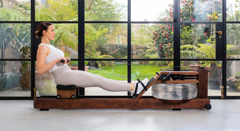 A woman in a white tank top and light grey leggings is rowing on a WaterRower Original wooden water row machine in front of a large black-framed window overlooking a green garden.