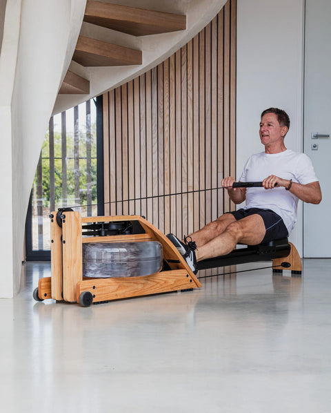 A man using a wooden WaterRower Hybrid water rowing machine in a modern indoor space with wood-panelled walls and a curved staircase.