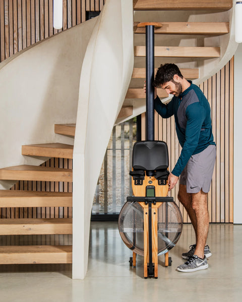 A man in athletic wear stands next to a WaterRower Hybrid on it's end in a modern indoor setting with a wooden staircase.