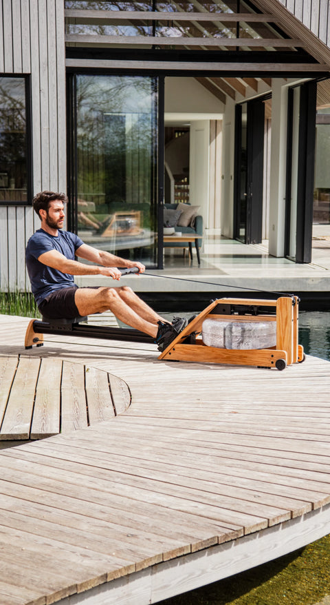 A man using a wooden WaterRower Hybrid rowing machine on an outdoor deck in front of a modern wooden house.