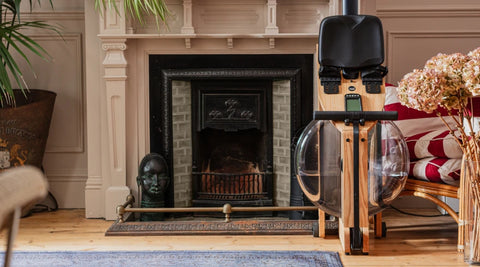 A wooden rowing machine with a black seat and clear water tank stands in a cozy, vintage-style room next to a fireplace.