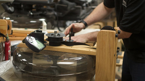A person in a workshop adjusts a dial indicator mounted on a wooden frame above a large clear WaterRower tank.