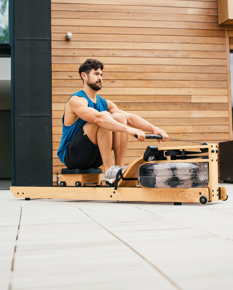A man sits on a WaterRower Lite wooden rowing machine outdoors in front of a wood-panelled wall, wearing a blue tank top and black shorts.