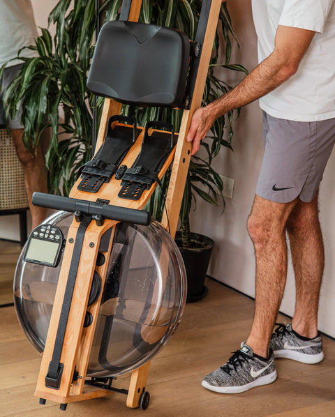 A WaterRower Lite wooden rowing machine with black seat and handle components, shown indoors with part of a person in a white shirt and grey Nike shorts touching the frame. A large green potted plant and wood floor are visible in the background.