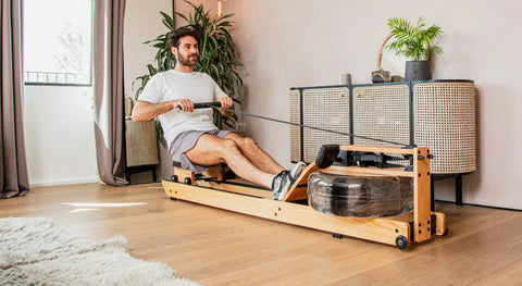 A man is using a WaterRower Lite wooden rowing machine in a bright, modern room with light wood floors, a white rug, and indoor plants.