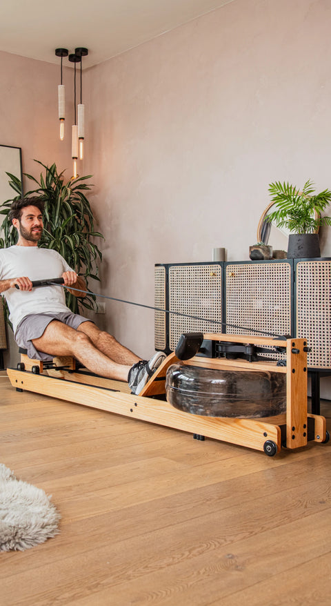 A man is using a WaterRower Lite wooden rowing machine in a bright, modern room with light wood floors, a white rug, and indoor plants.