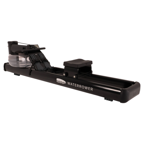 A black Waterrower rowing machine with a clear water tank and black footrest, shown from a three-quarters angle on a solid white background.